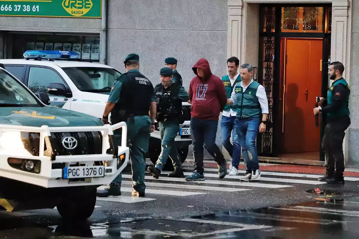 Varios agentes de la Guardia Civil escoltan a una persona con sudadera roja y capucha mientras lo sacan de un edificio hacia la calle, junto a vehículos oficiales estacionados. Varios agentes de la Guardia Civil escoltan a una persona con sudadera roja y capucha mientras lo sacan de un edificio hacia la calle, junto a vehículos oficiales estacionados.
