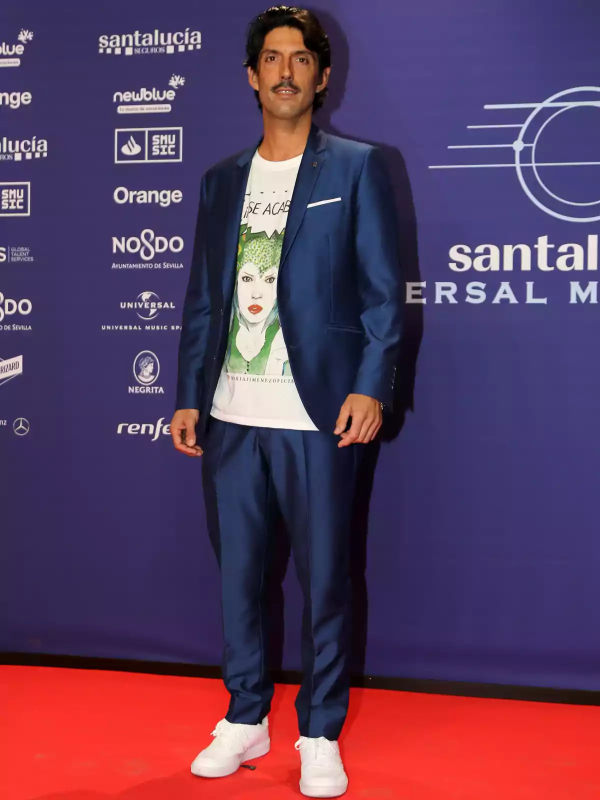 Hombre con traje azul y camiseta blanca con diseño gráfico, posando en una alfombra roja frente a un fondo con logotipos de patrocinadores.