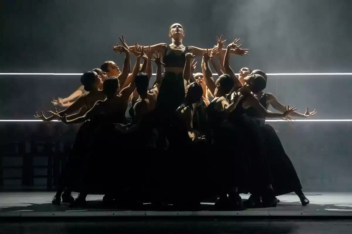 Grupo de bailarinas vestidas de negro realizando una coreografía dramática con una bailarina en el centro levantando los brazos mientras las demás la rodean y extienden sus manos hacia ella en un escenario con luces tenues y fondo oscuro Grupo de bailarinas vestidas de negro realizando una coreografía dramática con una bailarina en el centro levantando los brazos mientras las demás la rodean y extienden sus manos hacia ella en un escenario con luces tenues y fondo oscuro