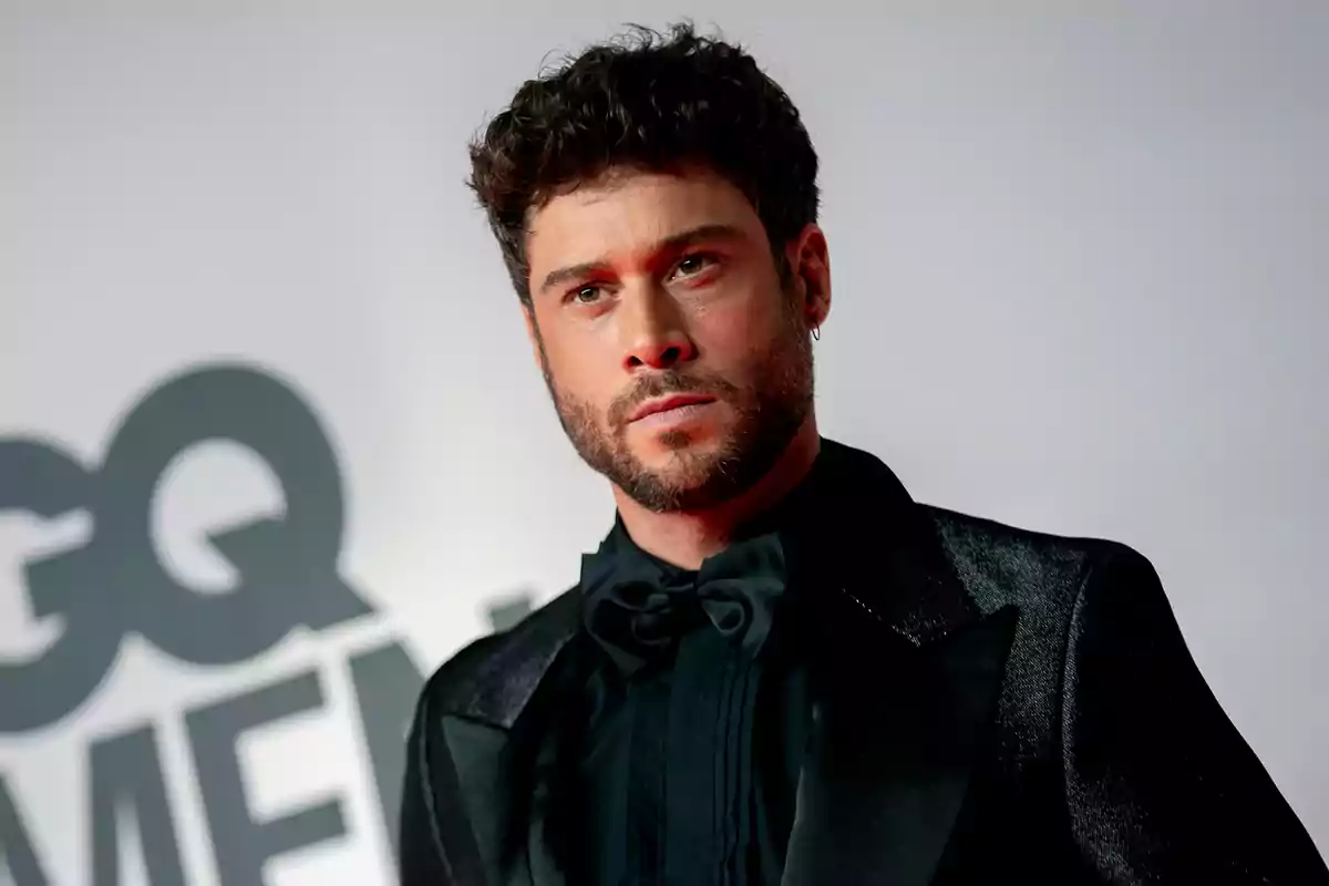 Hombre con barba y cabello rizado en un evento de gala con fondo de logotipo.