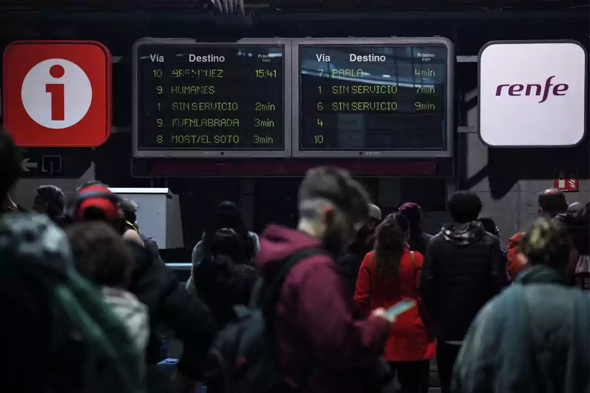 Personas esperando en una estación de tren con pantallas que muestran los destinos y tiempos de llegada de los próximos trenes.