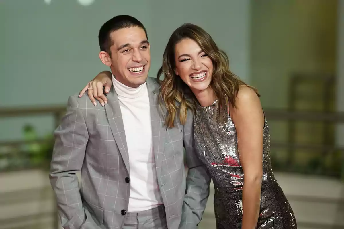 Dos personas sonrientes posan juntas en un evento formal, una lleva un traje gris y la otra un vestido brillante.