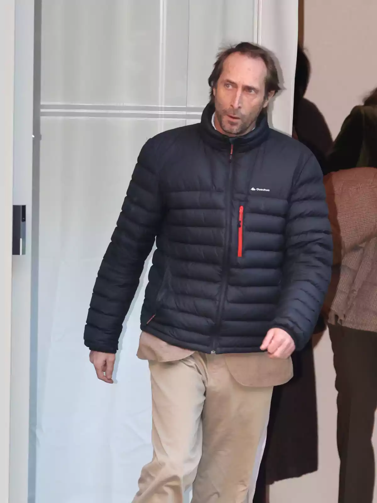 Un hombre con chaqueta acolchada negra y pantalones claros caminando por un pasillo. Un hombre con chaqueta acolchada negra y pantalones claros caminando por un pasillo.