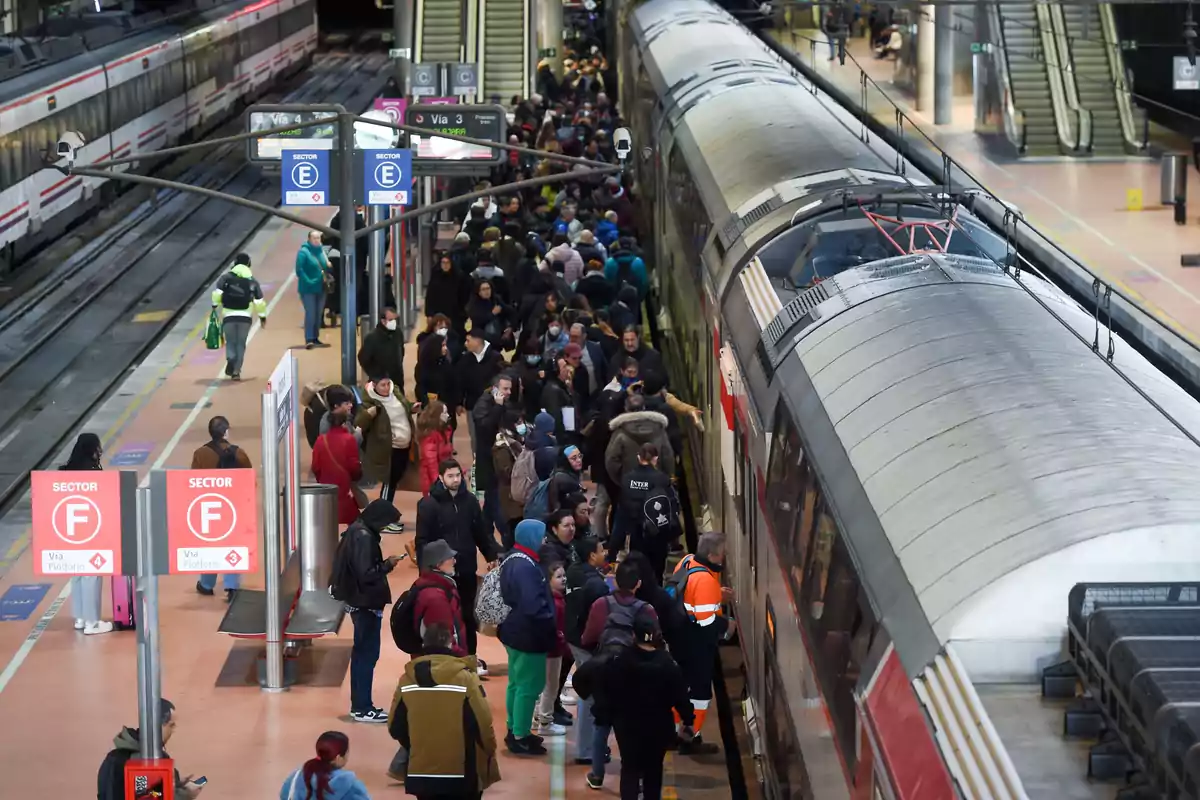 Una multitud de personas espera en el andén de una estación de tren, con varios trenes estacionados y señales de sector visibles.