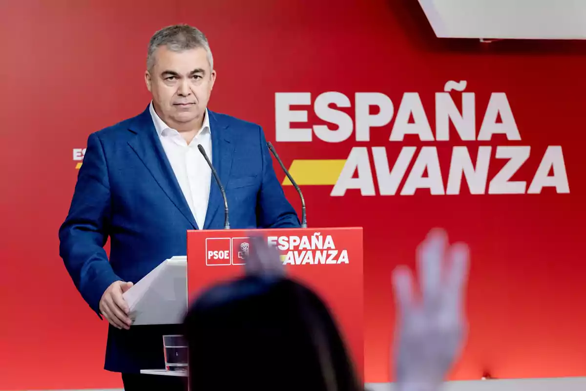 Hombre de cabello canoso con saco azul y camisa blanca hablando en un atril rojo con el logo del PSOE y el lema España Avanza sobre un fondo rojo Hombre de cabello canoso con saco azul y camisa blanca hablando en un atril rojo con el logo del PSOE y el lema España Avanza sobre un fondo rojo