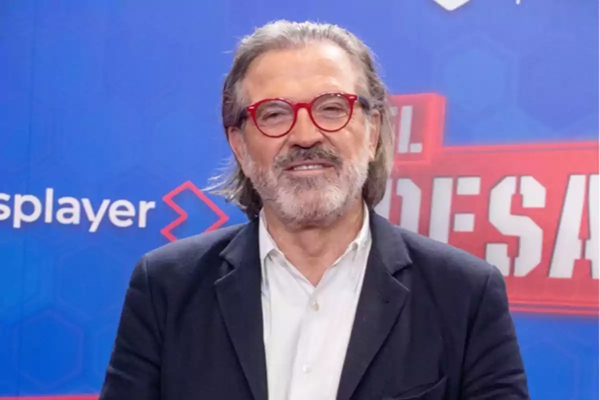 Un hombre con gafas rojas y barba posando frente a un fondo azul con texto.