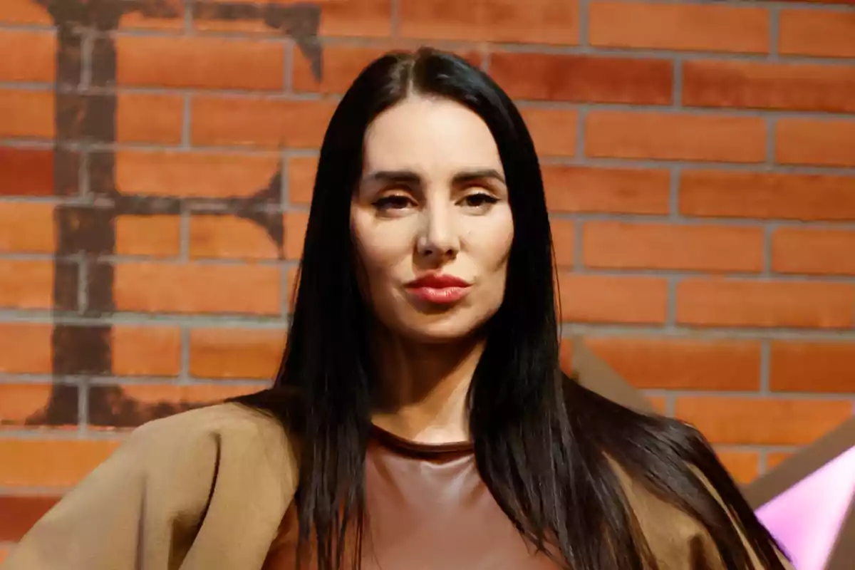 Mujer de cabello largo y oscuro con expresión seria posando frente a una pared de ladrillos