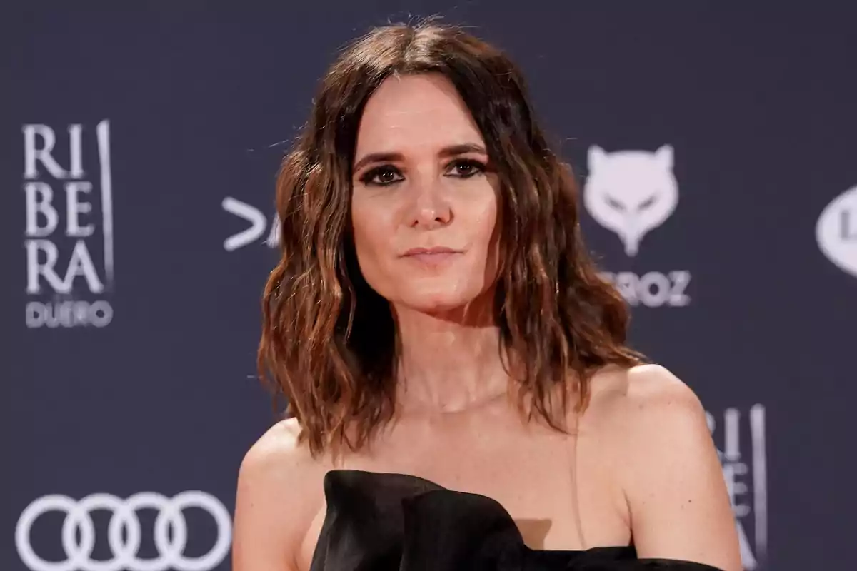 Una mujer con cabello ondulado y vestido negro posando en un evento con fondo de logotipos.