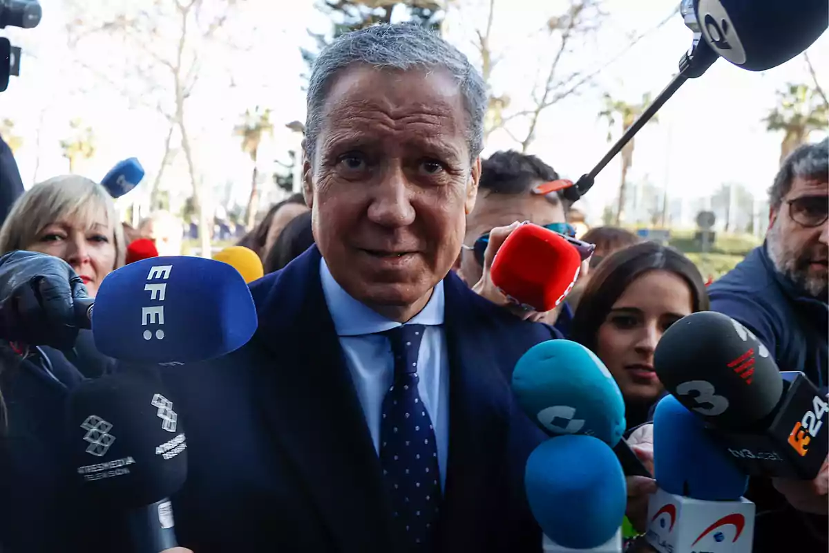 Un hombre de traje rodeado de micrófonos de diferentes medios de comunicación en un entorno al aire libre.