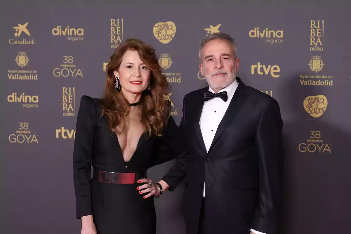 Una mujer y un hombre posan juntos en la alfombra roja de los Premios Goya, ambos vestidos de manera elegante; ella lleva un vestido negro con un cinturón metálico y él un esmoquin negro con pajarita.