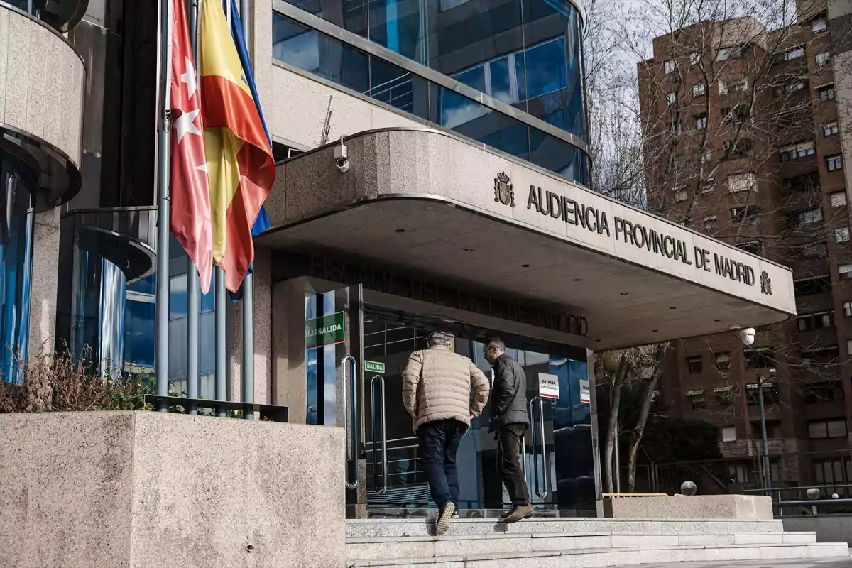 Dos personas suben las escaleras hacia la entrada de la Audiencia Provincial de Madrid, con banderas ondeando a un lado y edificios residenciales al fondo. Dos personas suben las escaleras hacia la entrada de la Audiencia Provincial de Madrid, con banderas ondeando a un lado y edificios residenciales al fondo.