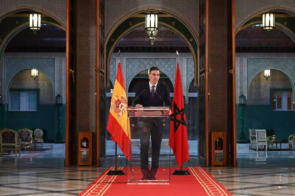 Un hombre de pie detrás de un podio con banderas de España y Marruecos en un entorno arquitectónico elegante.