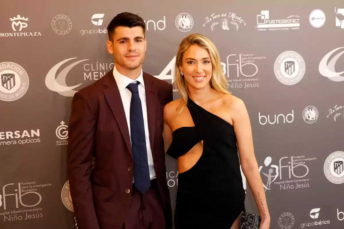 Una pareja posando en un evento formal con un fondo de logotipos de patrocinadores.