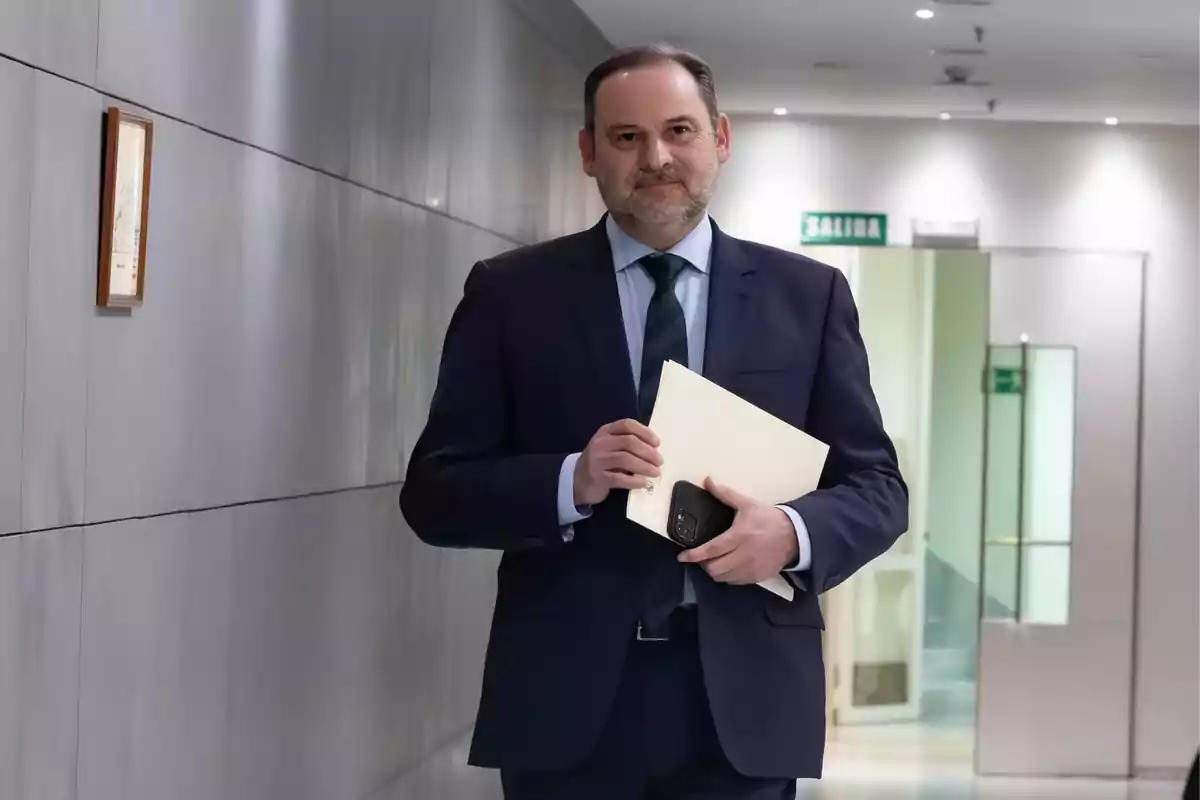 Hombre de traje caminando por un pasillo con una carpeta y un teléfono en la mano.