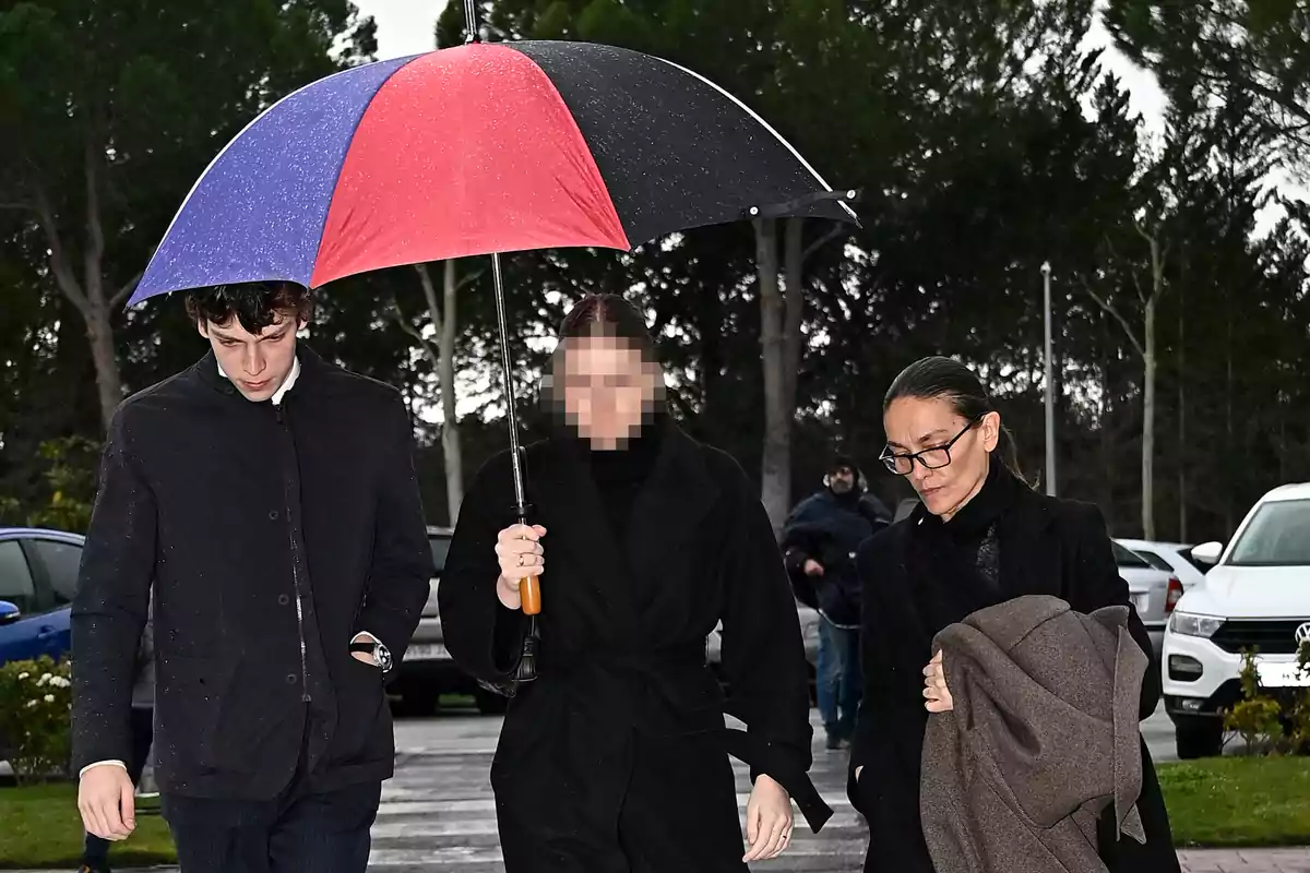 Tres personas caminando bajo la lluvia con un paraguas de colores en un entorno exterior con árboles y coches estacionados. Tres personas caminando bajo la lluvia con un paraguas de colores en un entorno exterior con árboles y coches estacionados.