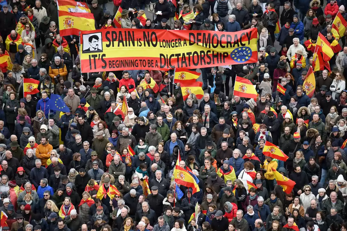 Una multitud de personas se ha reunido en una manifestación, muchas de ellas portando banderas de España y algunas de la Unión Europea, mientras sostienen una pancarta que dice "Spain is no longer a democracy, it's beginning to be a dictatorship".