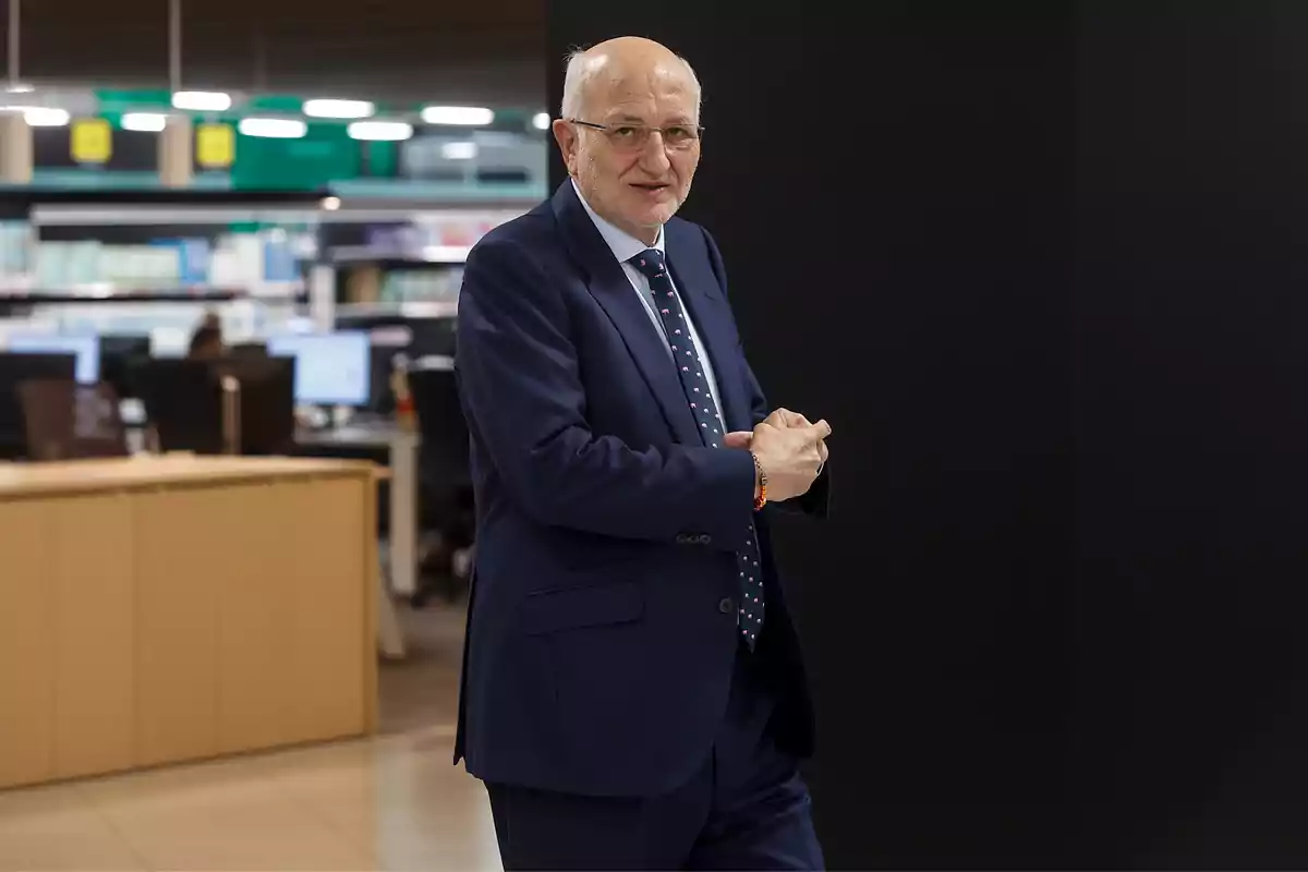 Un hombre mayor con traje azul y corbata de lunares en una oficina moderna con escritorios y computadoras al fondo.