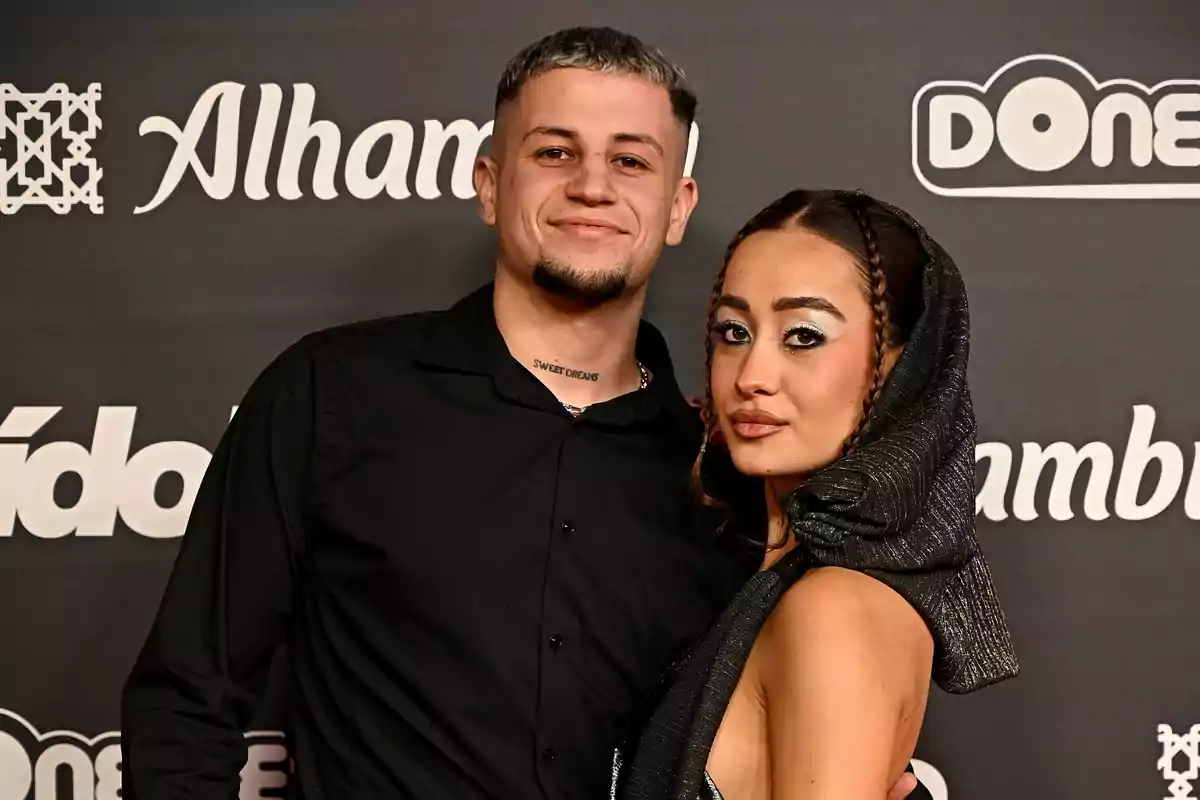 Una pareja posando en un evento con fondo de logotipos.