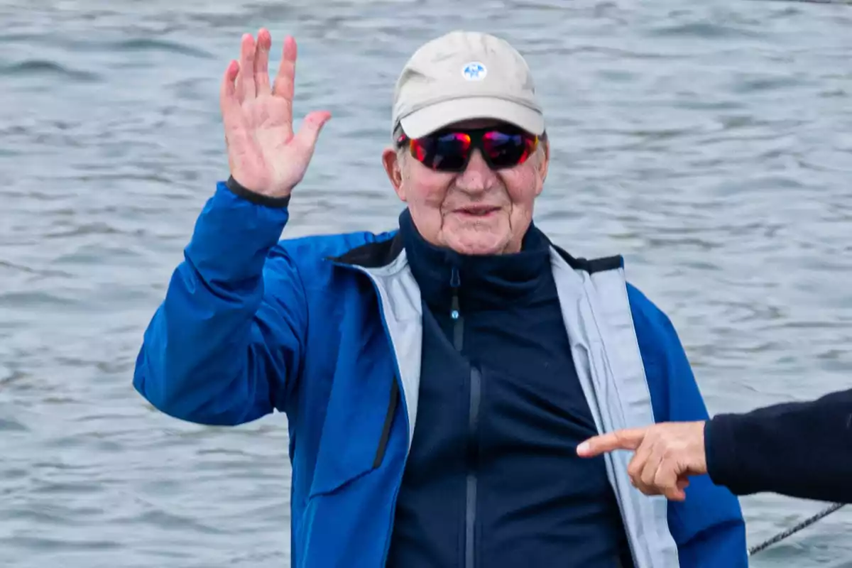 Un hombre con gorra y gafas de sol saluda con la mano cerca del agua.