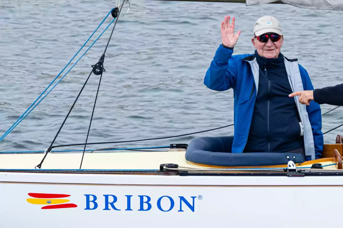 Un hombre con gorra y gafas de sol saluda desde un barco llamado Bribon en el agua.