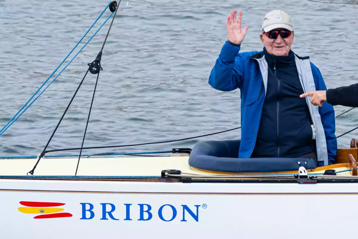 Hombre mayor con gorra y gafas de sol saludando desde un velero llamado Bribon en el agua Hombre mayor con gorra y gafas de sol saludando desde un velero llamado Bribon en el agua