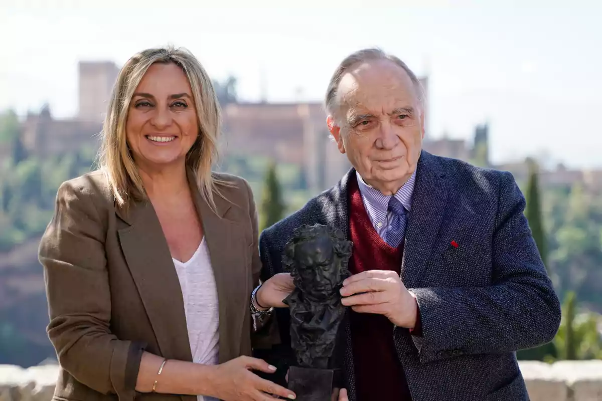 Dos personas sonrientes sosteniendo un busto en un entorno al aire libre con un edificio histórico de fondo.