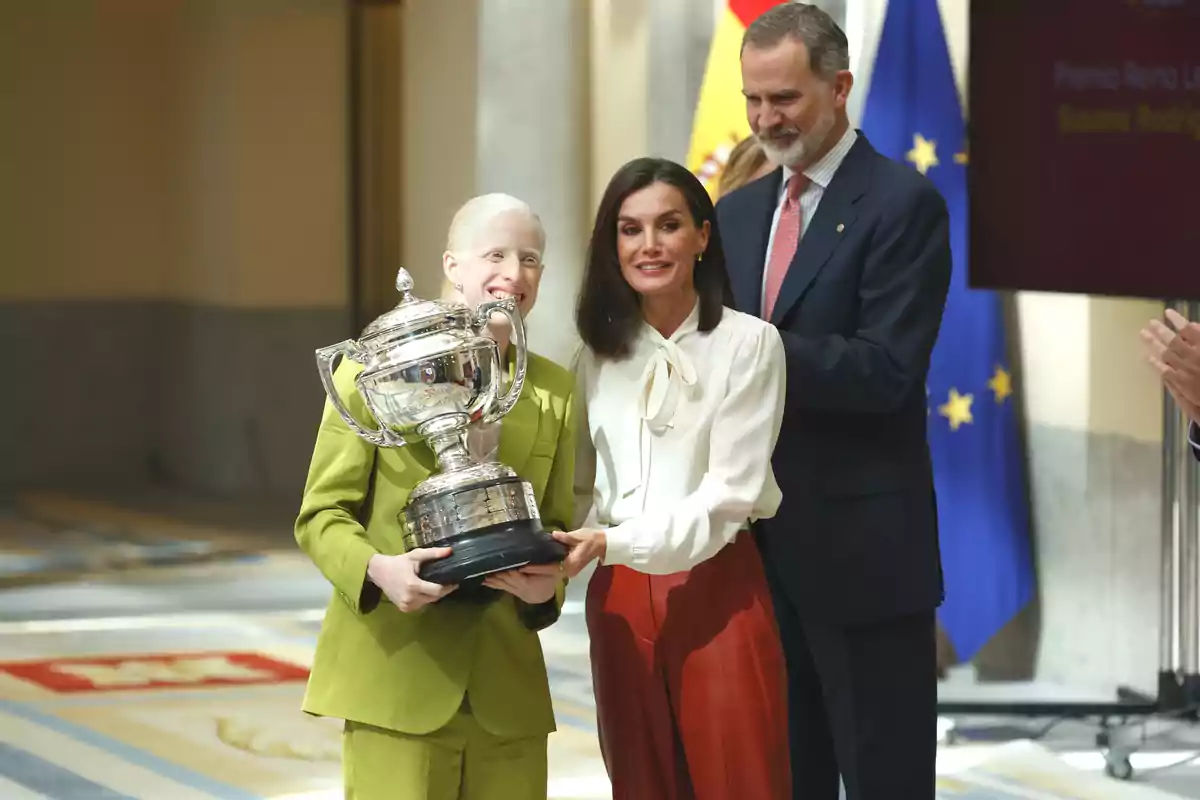 Tres personas posan con un gran trofeo plateado en un evento formal con banderas de fondo.