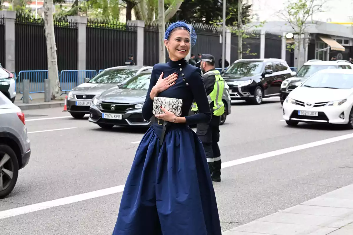Una mujer con un vestido azul oscuro y un tocado sonríe mientras camina por una calle con tráfico y policías en el fondo.