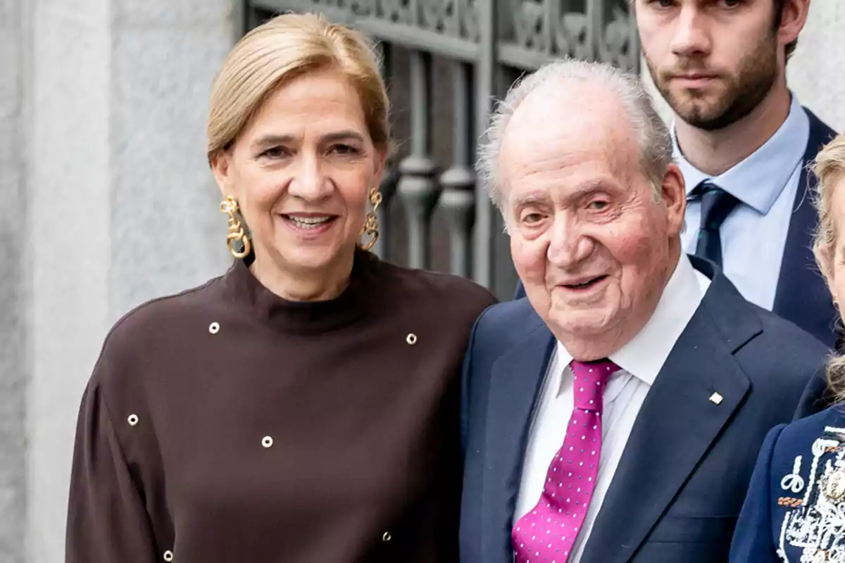 Una mujer de cabello rubio y un hombre mayor con traje y corbata fucsia posan juntos y sonríen frente a una reja de metal Una mujer de cabello rubio y un hombre mayor con traje y corbata fucsia posan juntos y sonríen frente a una reja de metal