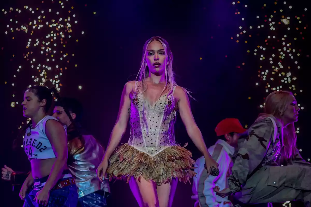 Una artista en el escenario con un vestido brillante rodeada de bailarines y luces de fondo.