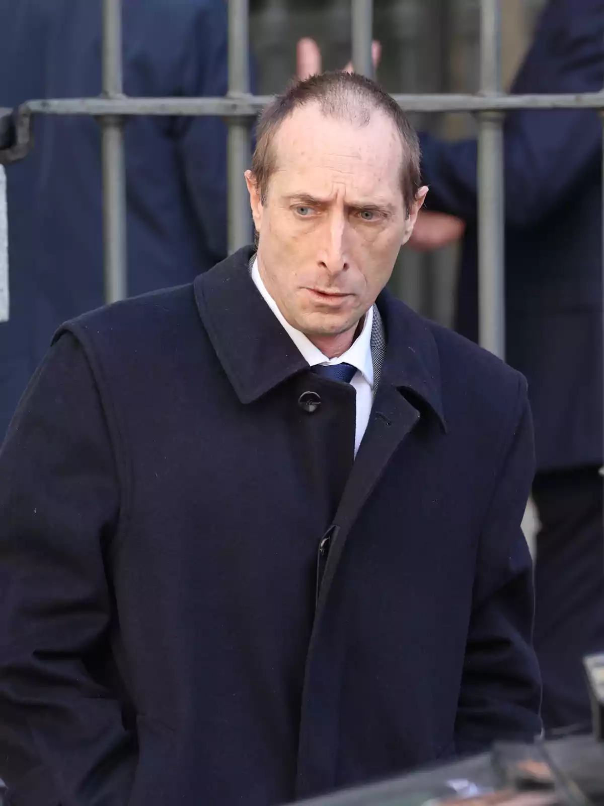 Un hombre con abrigo oscuro y expresión seria está de pie frente a una reja. Un hombre con abrigo oscuro y expresión seria está de pie frente a una reja.