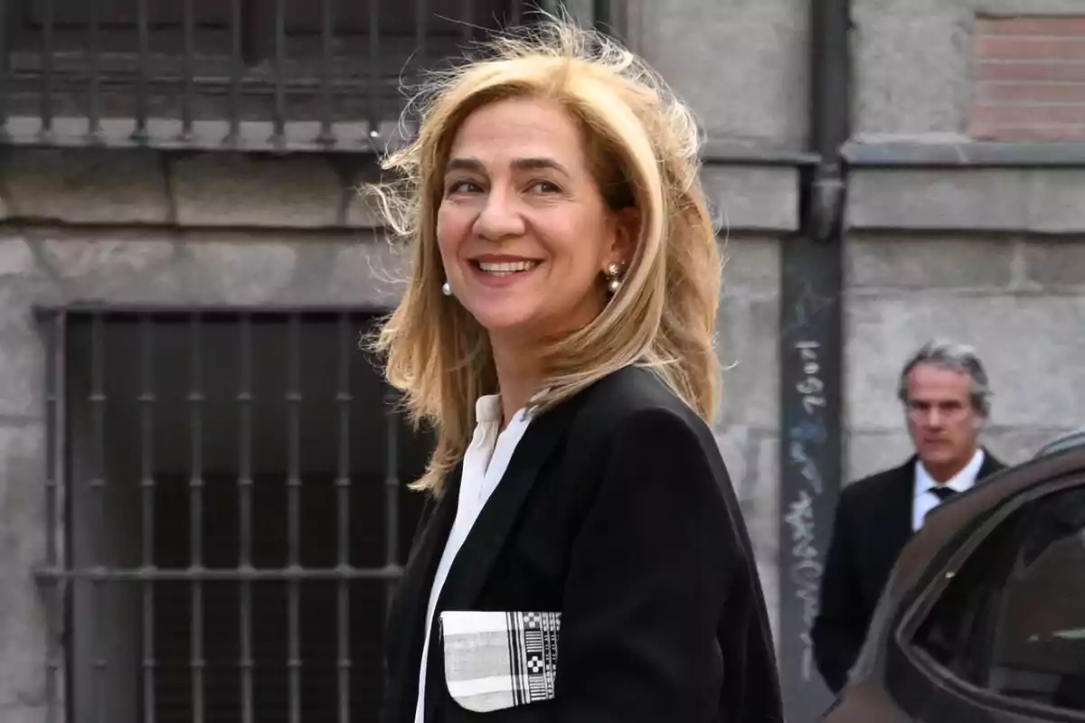 Mujer rubia sonriente con chaqueta negra y blusa blanca caminando en la calle frente a un edificio de piedra