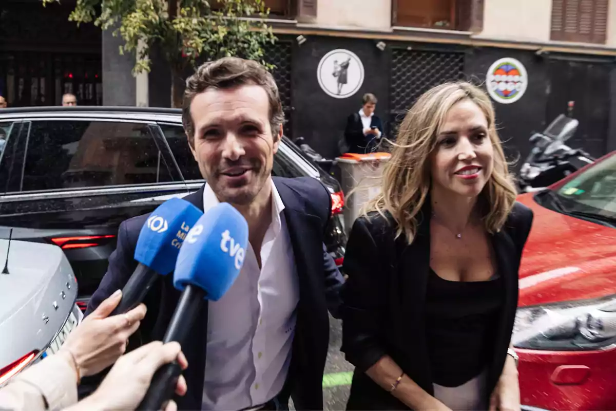 Una pareja sonriente es entrevistada por varios medios de comunicación en la calle, con micrófonos de diferentes cadenas de televisión frente a ellos.