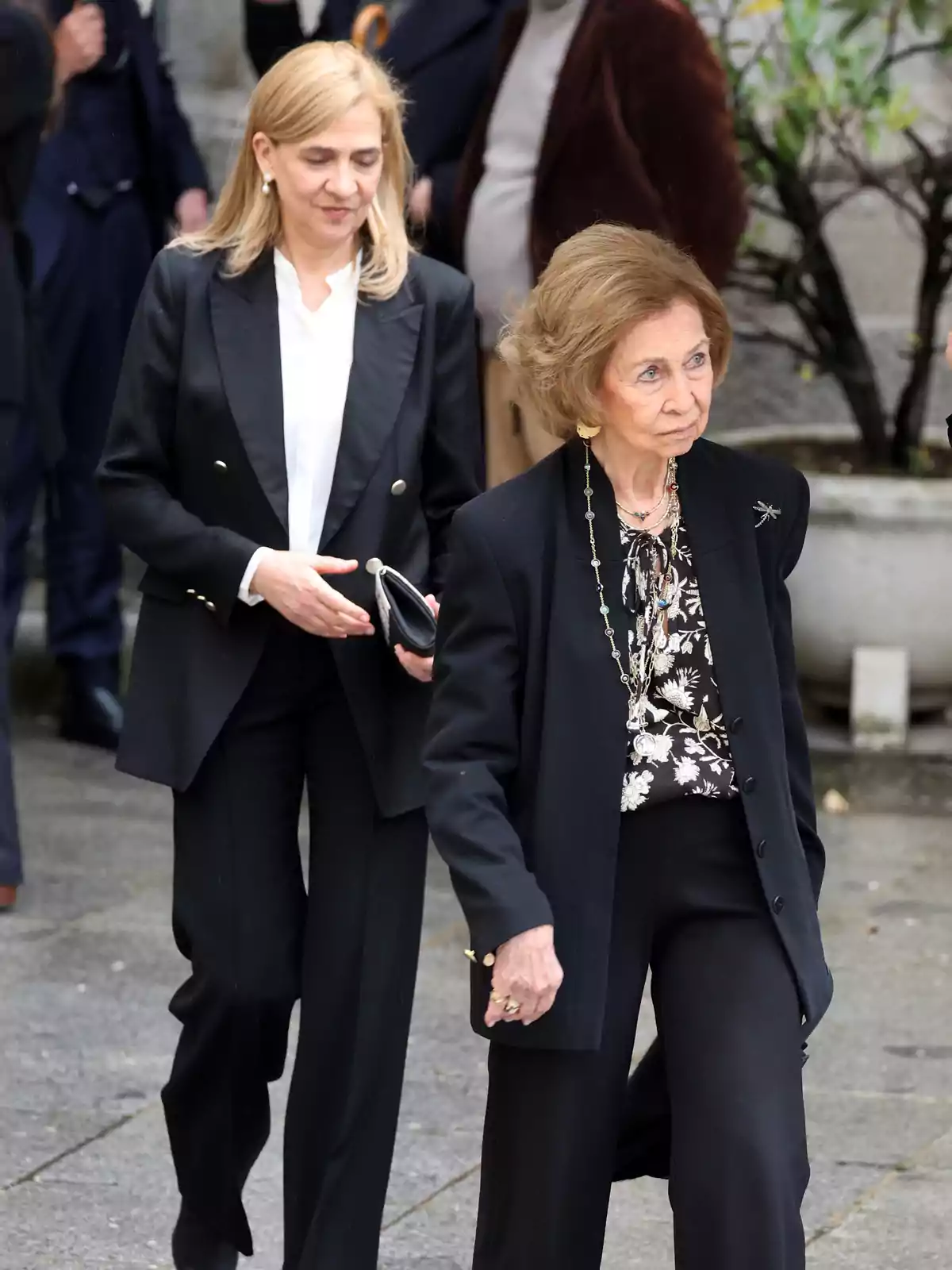 Dos mujeres vestidas de negro caminan al aire libre en un entorno urbano, una de ellas lleva un bolso y la otra un collar largo Dos mujeres vestidas de negro caminan al aire libre en un entorno urbano, una de ellas lleva un bolso y la otra un collar largo