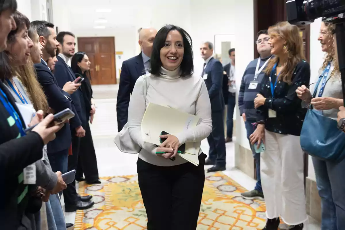 Una mujer sonriente camina por un pasillo rodeada de personas que la observan y sostienen teléfonos móviles.