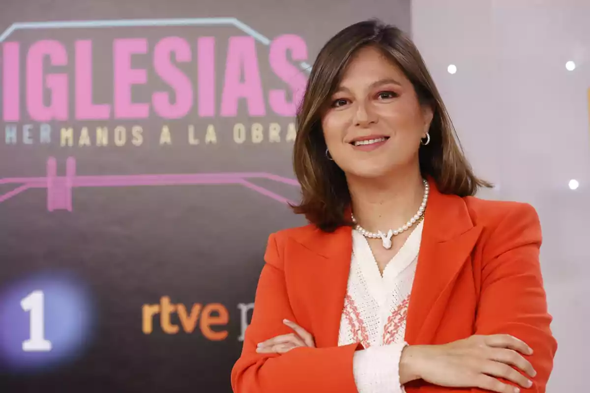 Una mujer con chaqueta naranja y collar de perlas sonríe frente a un cartel que dice "IGLESIAS HERMANOS A LA OBRA" y tiene los logotipos de RTVE y La 1.