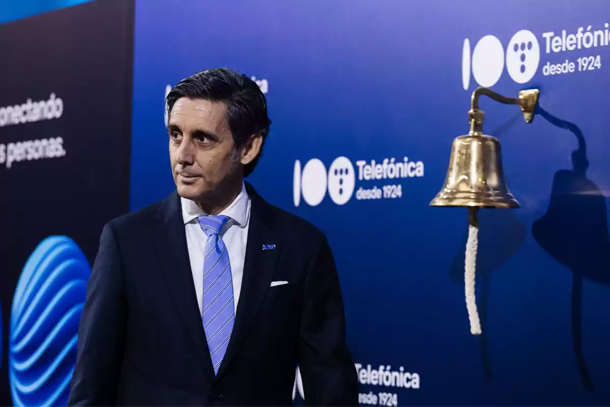 Un hombre de traje y corbata azul claro está de pie frente a un fondo azul con el logotipo de Telefónica y una campana dorada.