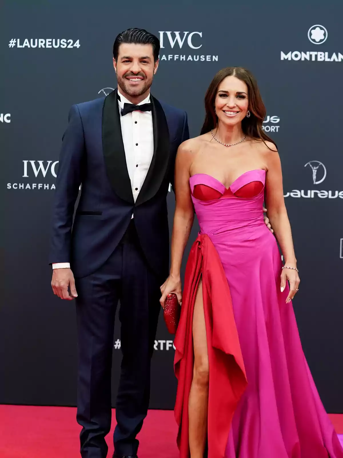 Pareja posando en una alfombra roja, él lleva un esmoquin azul marino con pajarita negra y ella un vestido largo rosa con abertura y detalles rojos, ambos sonríen frente a un fondo con logotipos de marcas. Pareja posando en una alfombra roja, él lleva un esmoquin azul marino con pajarita negra y ella un vestido largo rosa con abertura y detalles rojos, ambos sonríen frente a un fondo con logotipos de marcas.