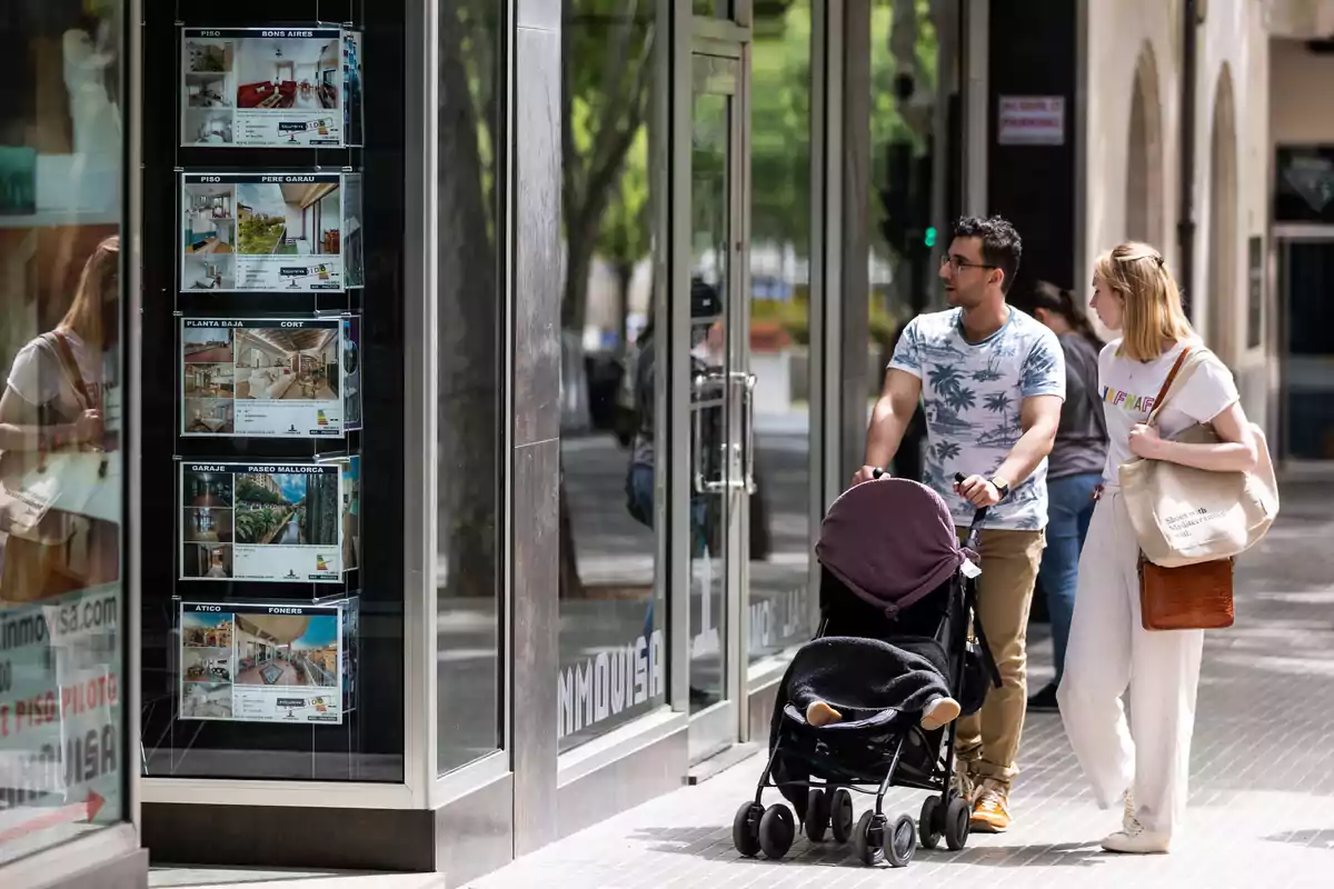 Una pareja con un cochecito de bebé observa anuncios de propiedades en la vitrina de una inmobiliaria.