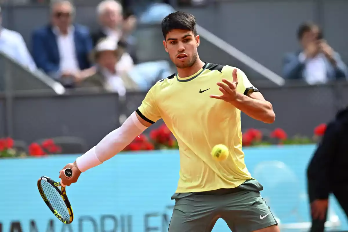 Un jugador de tenis con camiseta amarilla y pantalones cortos grises se prepara para golpear una pelota con su raqueta en un partido, mientras el público observa desde las gradas.