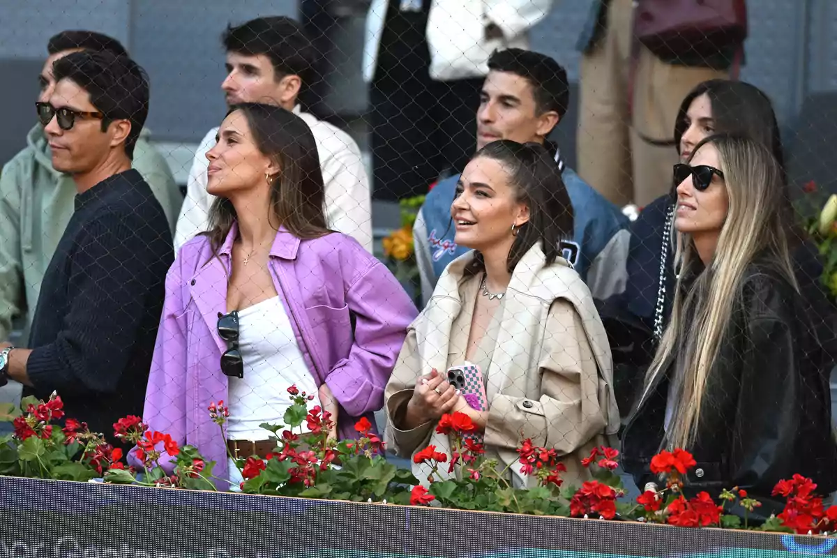 Un grupo de personas observando un evento deportivo detrás de una red, con flores rojas en primer plano.