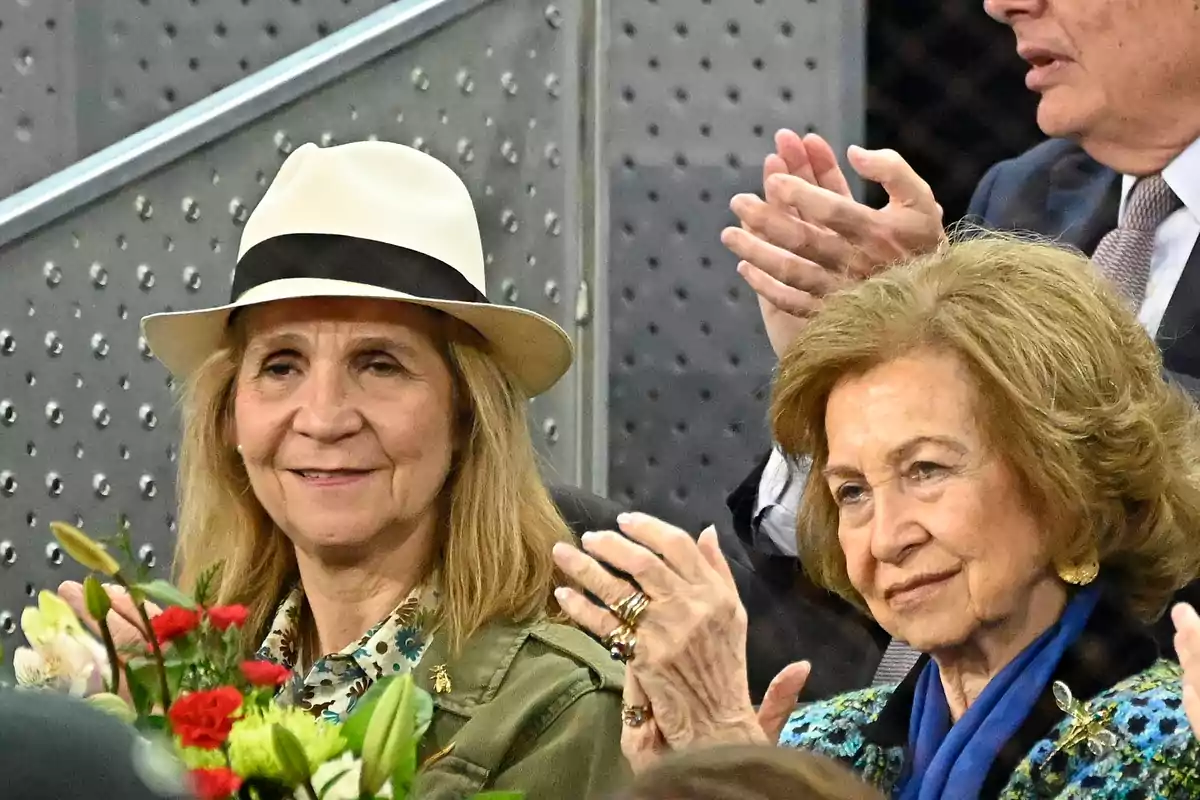 Dos mujeres sentadas en un evento, una lleva un sombrero blanco y la otra una bufanda azul, ambas parecen estar disfrutando del momento.