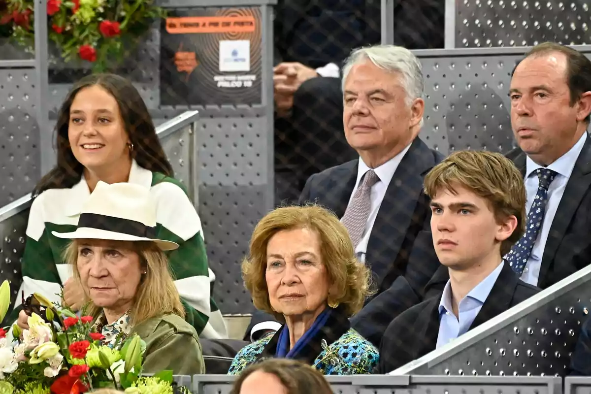 Un grupo de personas está sentado en las gradas de un evento deportivo, algunas de ellas parecen estar observando con atención mientras otras conversan o sonríen.