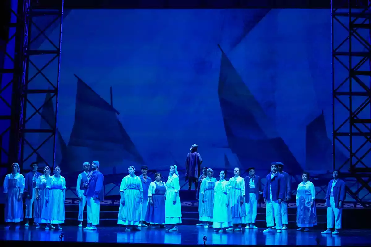 Un grupo de personas vestidas con ropa de época se encuentra en el escenario de un teatro con una escenografía azul que simula velas de barcos al fondo Un grupo de personas vestidas con ropa de época se encuentra en el escenario de un teatro con una escenografía azul que simula velas de barcos al fondo