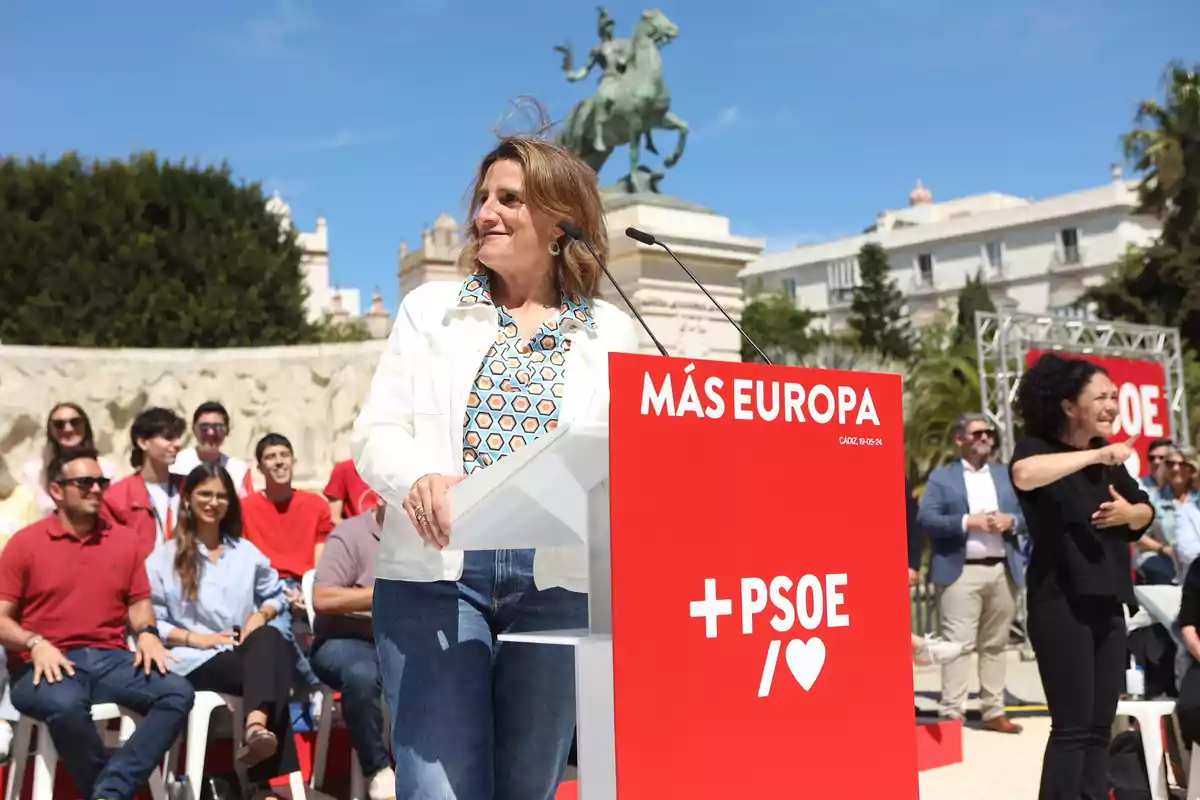 Una mujer en un evento al aire libre hablando en un podio con el logotipo del PSOE y el texto 