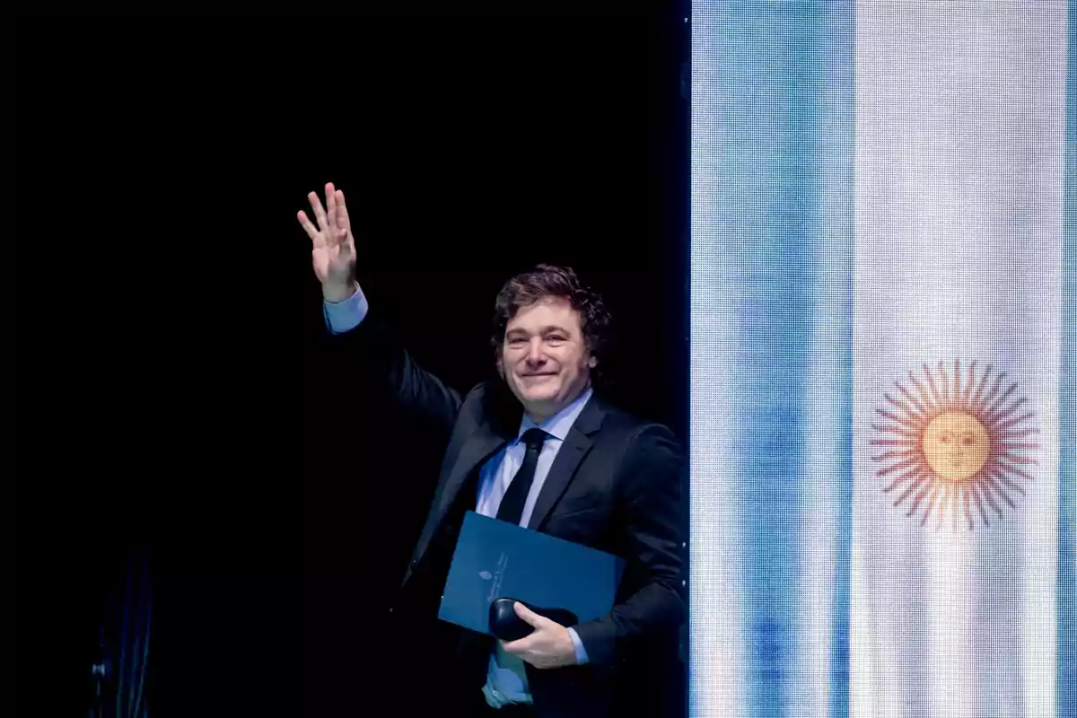 Persona con traje oscuro y corbata negra saludando con la mano levantada, sosteniendo un documento azul, con una bandera de Argentina de fondo.