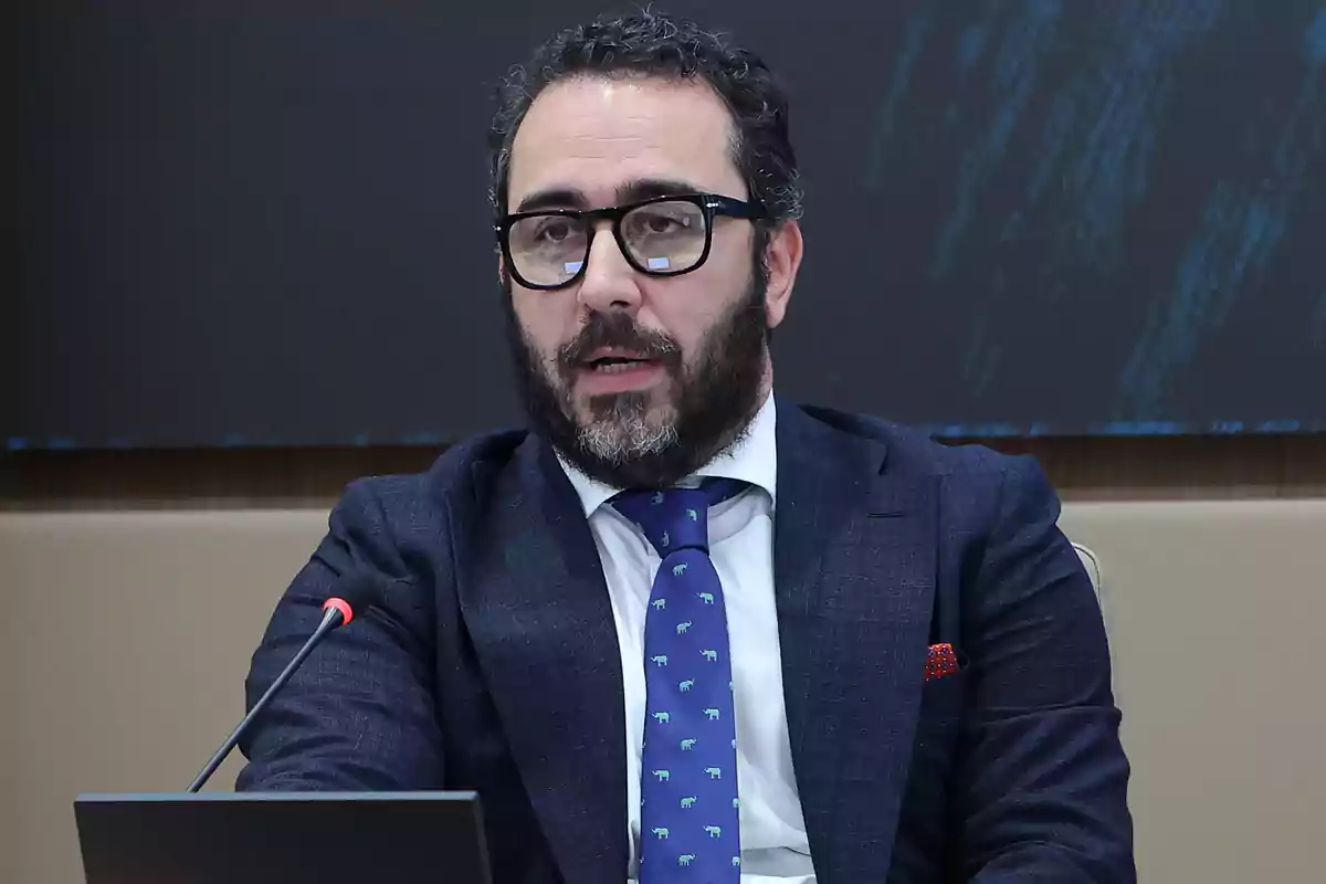 Hombre con barba y gafas hablando en un micrófono durante una conferencia.