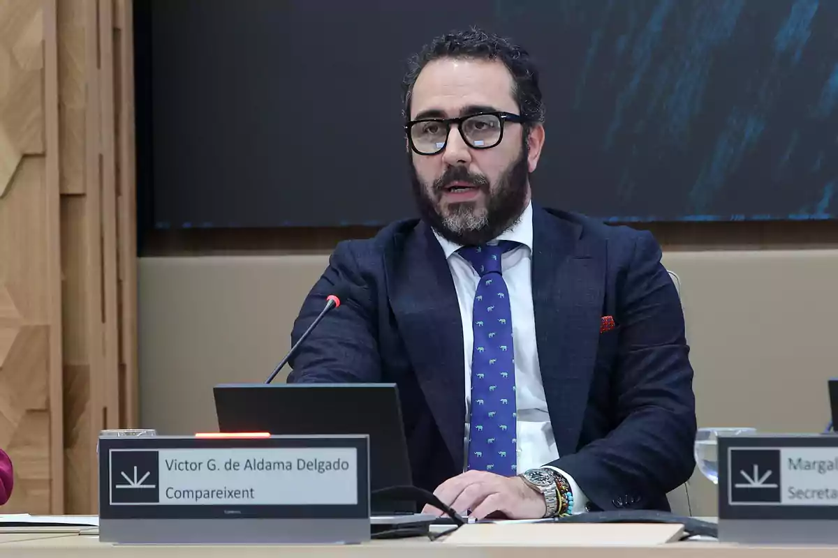 Un hombre con barba y gafas está sentado en una mesa de conferencias, hablando frente a un micrófono y una placa con su nombre.