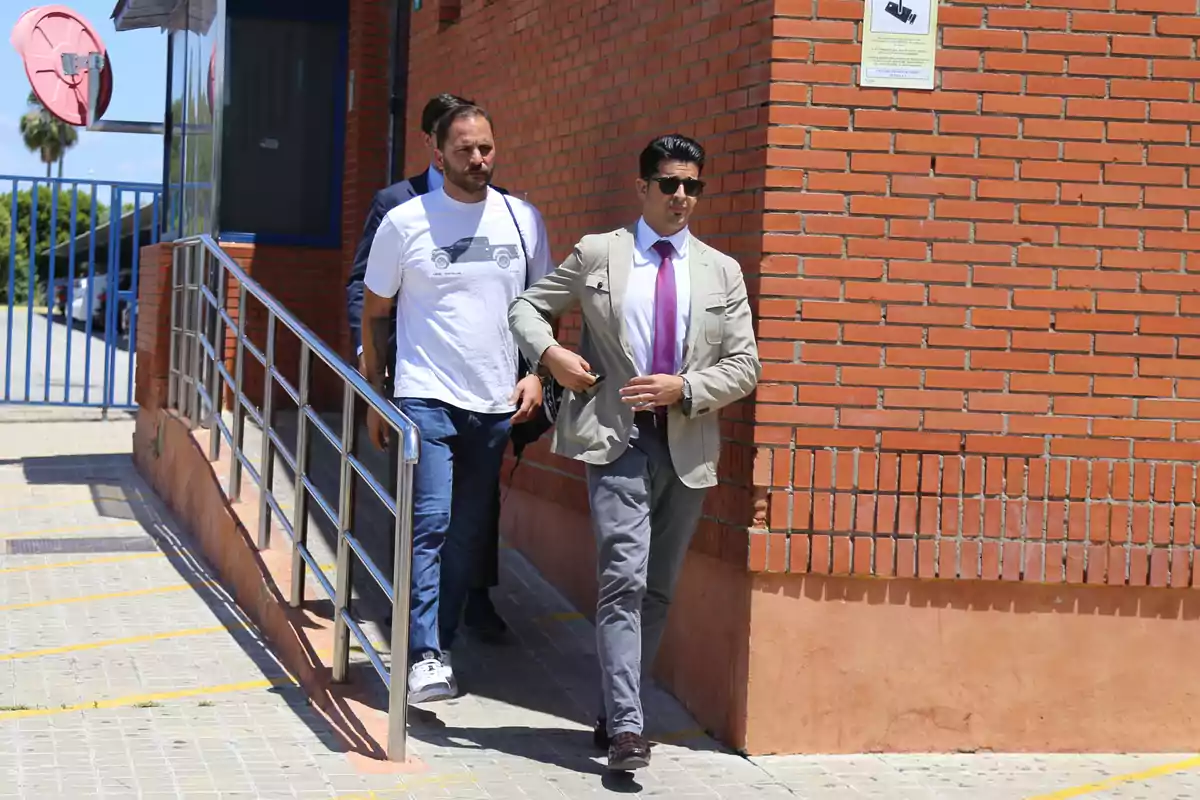 Dos hombres caminan por una acera junto a un edificio de ladrillo rojo, uno de ellos lleva una camiseta blanca y jeans, mientras que el otro viste un traje claro con corbata morada. Dos hombres caminan por una acera junto a un edificio de ladrillo rojo, uno de ellos lleva una camiseta blanca y jeans, mientras que el otro viste un traje claro con corbata morada.