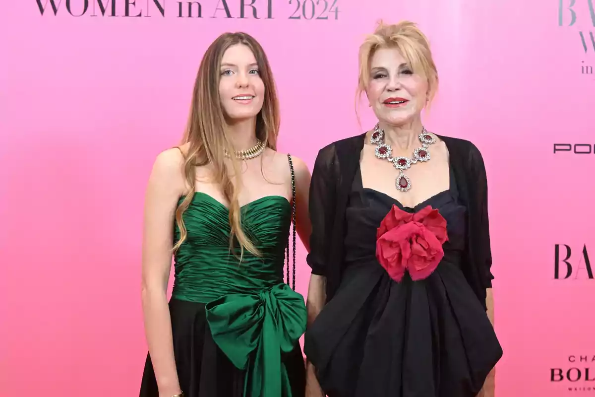 Dos mujeres posan en un evento de arte, una con un vestido verde y la otra con un vestido negro con un adorno rojo, ambas con joyería elegante y un fondo rosa con texto.
